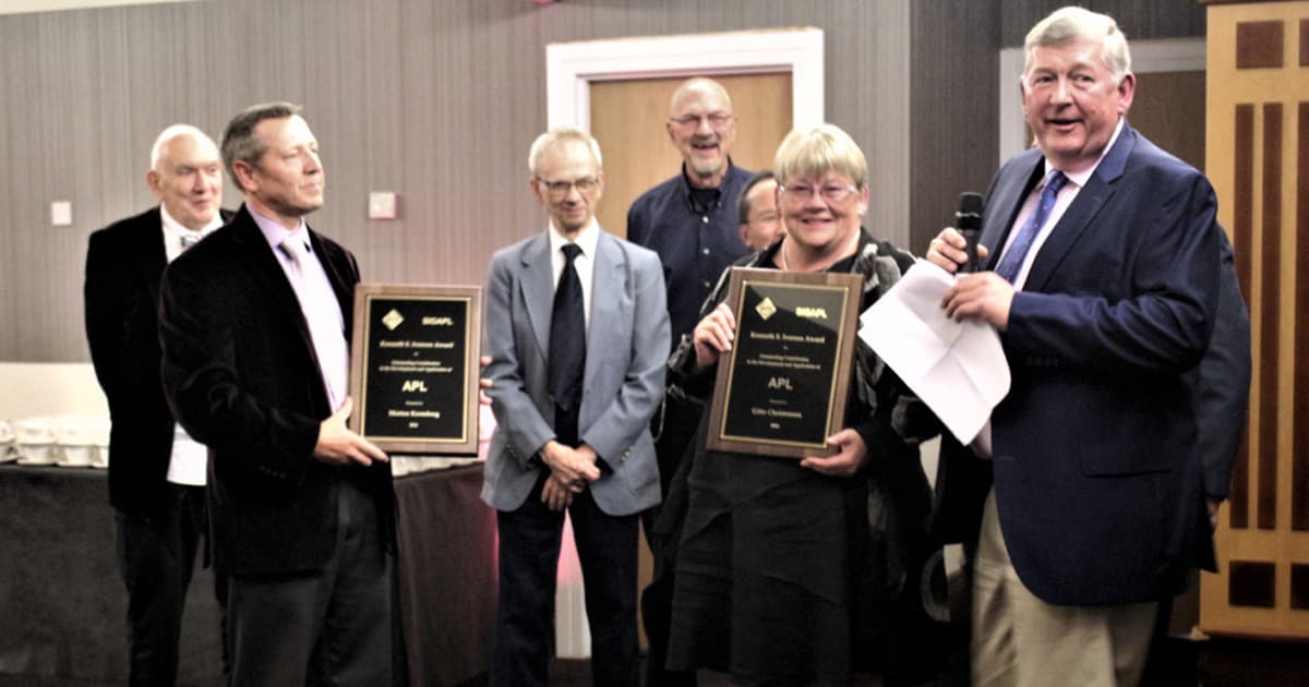 Morten Kromberg and Gitte Christensen receive the 2016 Iverson Award at a formal event. Gitte holds a plaque while Paul Grosvenor of Optima Systems Ltd speaks into a microphone.