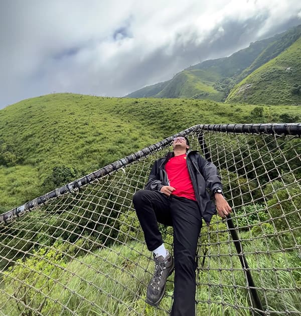 Aarush Bhat relaxing on a rope net suspended above lush green hills under a cloudy sky.