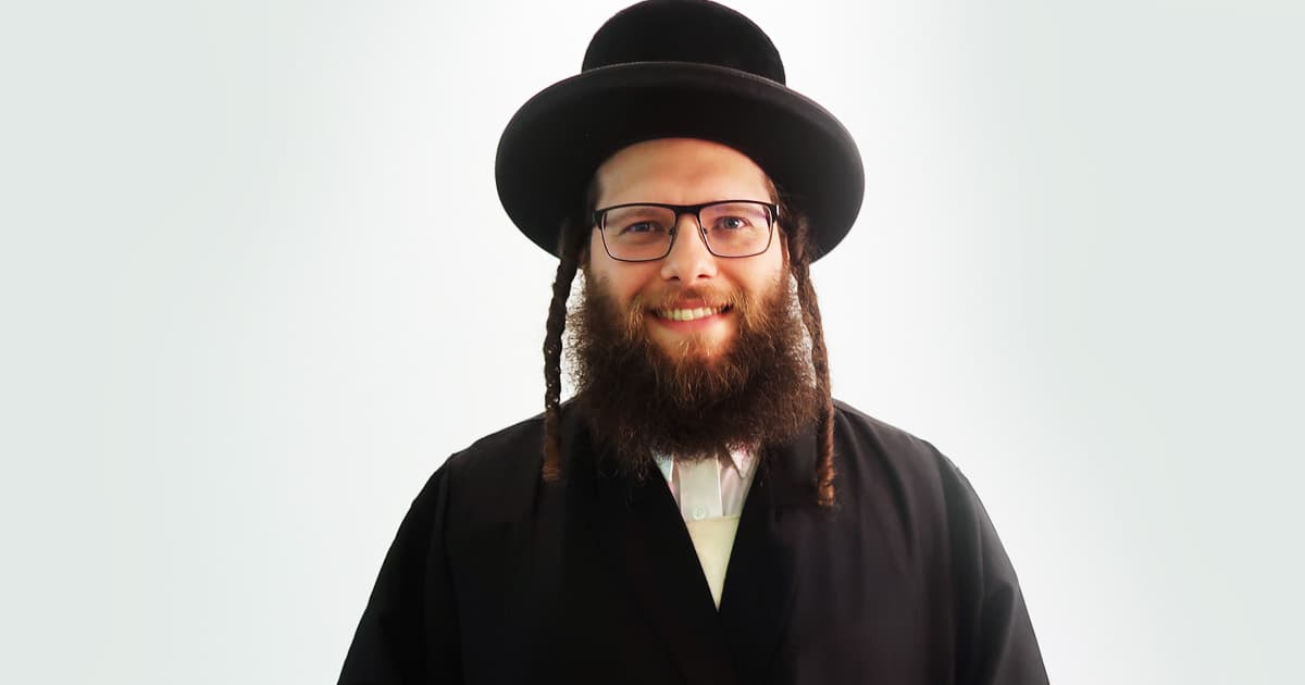 Adám Brudzewsky smiles at the camera, wearing traditional black attire and a wide-brimmed hat, with sidelocks and a full beard.