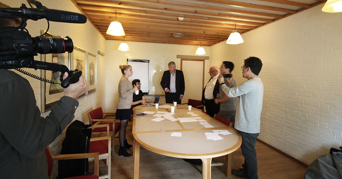 A behind-the-scenes view at Dyalog ’19 showing a camera crew filming a group in a small meeting room at Konventum. Several attendees are gathered around a table with notes and coffee cups as short films about APL are recorded under warm lighting and a wooden ceiling.