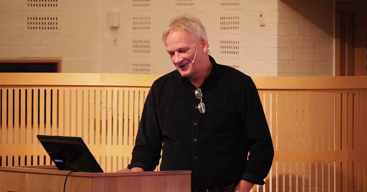 Tomas Gustafsson stands at a podium delivering a presentation at Dyalog ’19. He is smiling, wearing a dark shirt with glasses hanging from the collar, and a headset microphone. A wooden screen and light brick wall are visible in the background of the conference room.