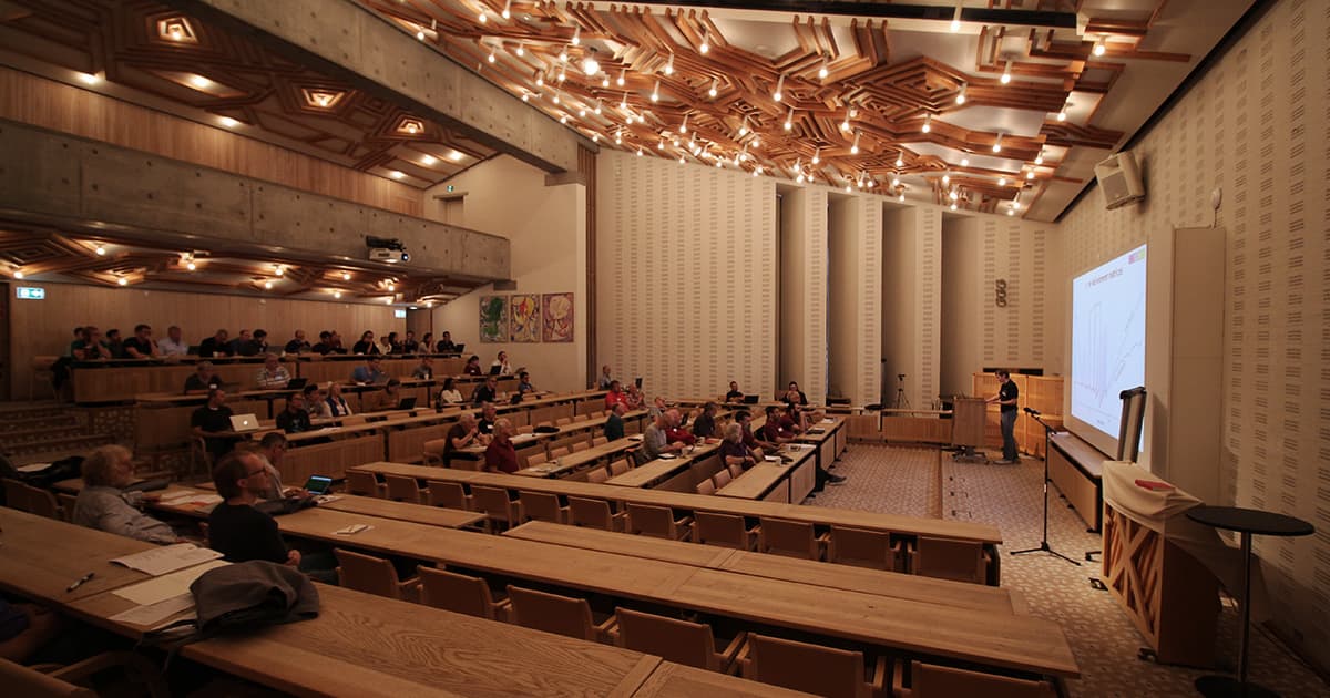A speaker presents a slide to an audience in the main lecture hall at Konventum during Dyalog ’19. The room features tiered wooden seating, abstract wooden ceiling panels with built-in lighting, and a large projection screen on the right. Attendees are seated throughout the hall, listening and taking notes.