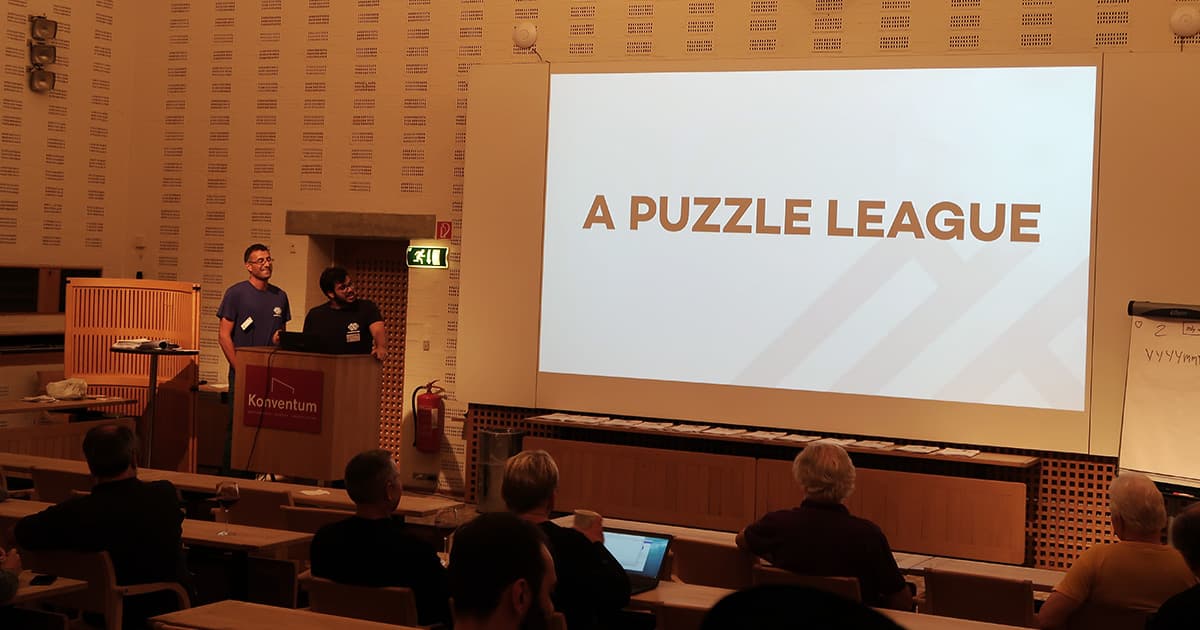 Roberto Minelli and Pietro Bulleri stand at a podium in a lecture hall at Konventum, presenting to an audience. A large screen behind them displays the title “A Puzzle League.” Attendees are seated in rows, watching the talk, with a whiteboard showing handwritten notes on the right-hand side.
