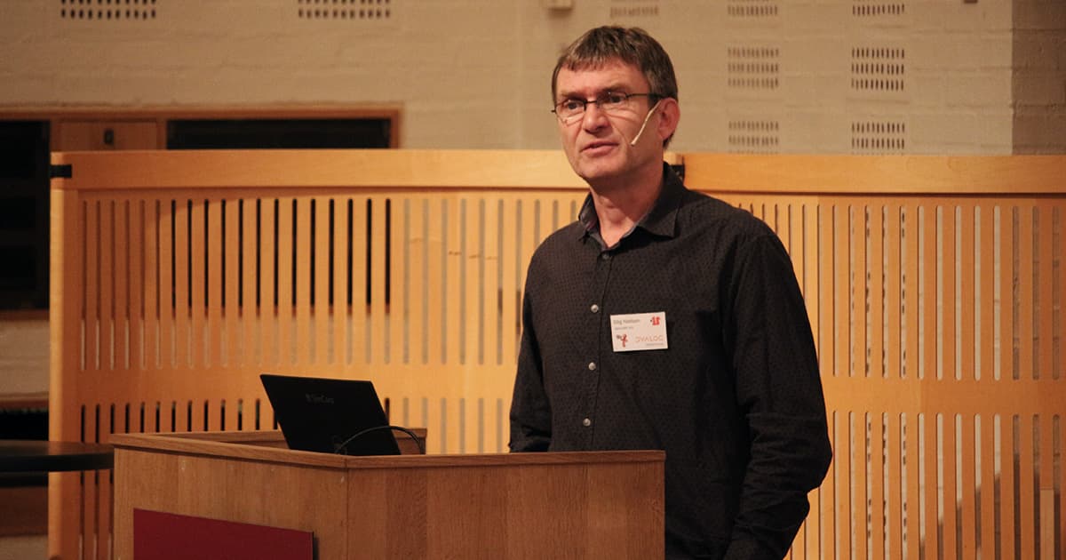 Stig Nielsen of SimCorp stands at the podium during Dyalog ’19, wearing a headset microphone and a name badge. He appears mid-presentation, with a laptop in front of him and a wood-panelled backdrop behind.
