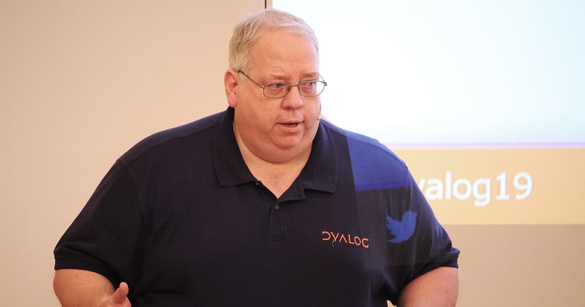 Brian Becker of Dyalog Ltd stands in front of a presentation screen at Dyalog ’19, wearing a Dyalog-branded navy polo shirt and headset microphone, introducing the HTMLRenderer tool.