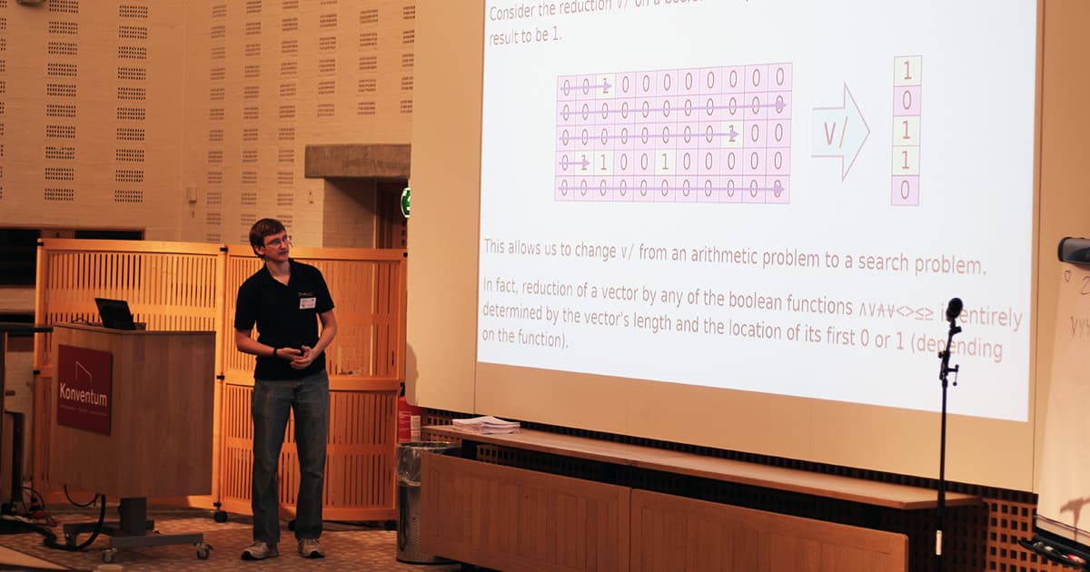 Marshall Lochbaum stands beside a large screen during his presentation at Dyalog ’19, showing a diagram of Boolean reductions and APL expressions. He gestures toward the projection, which includes visual representations of vectors and symbols like ∨. A Konventum podium and microphone stand are visible nearby.