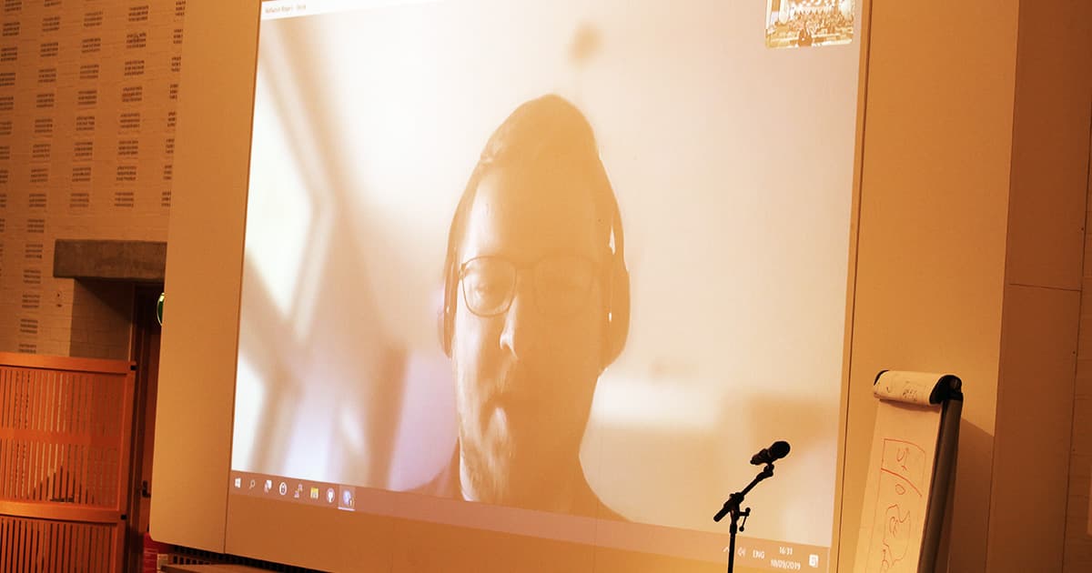 A large projected screen shows Nathan Rogers speaking via video call during Dyalog ’19. He is wearing glasses and a headset, presenting remotely from Colorado.