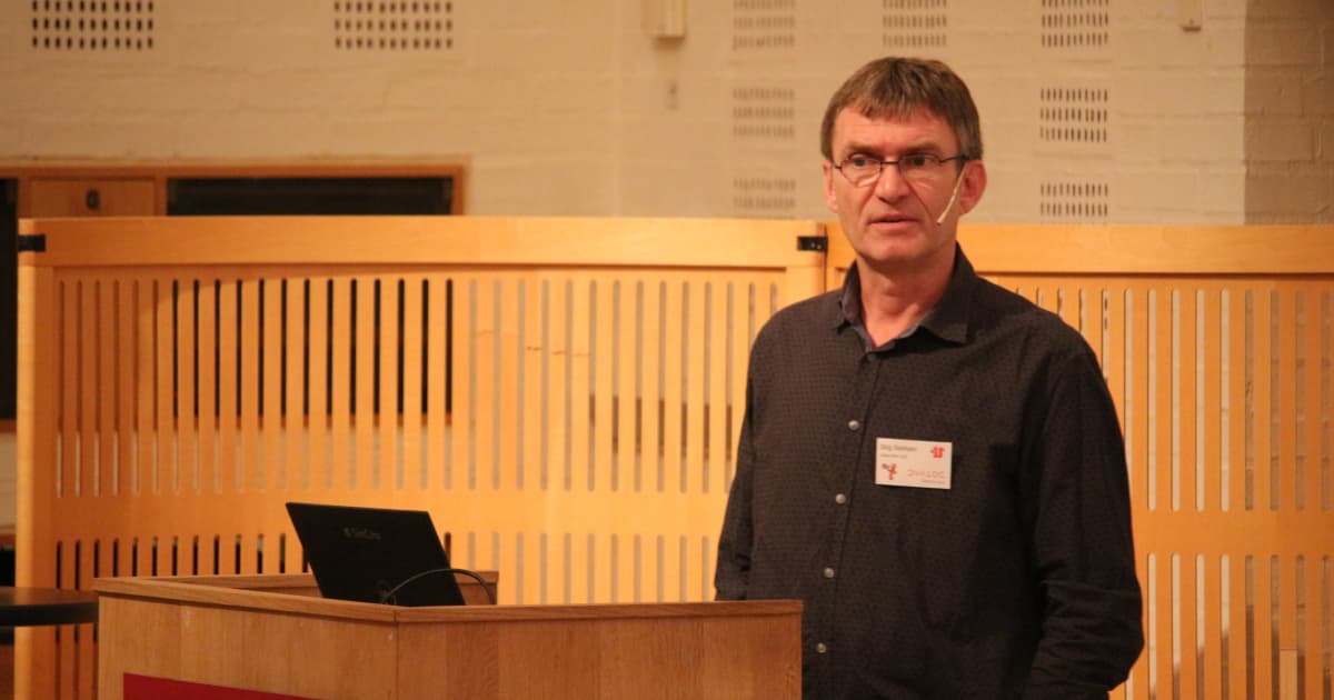 Stig Nielsen presenting at Dyalog ’19 in Elsinore. He stands at a wooden podium with a laptop, wearing a dark shirt and glasses, speaking into a headset microphone.
