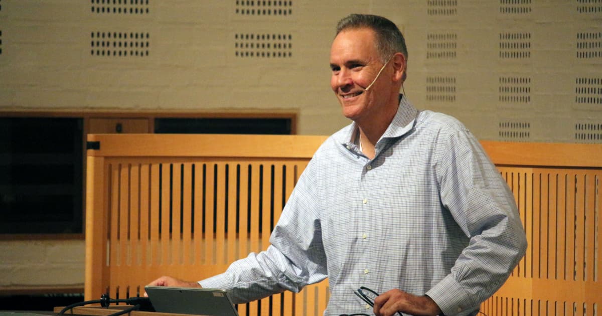 Paul Mansour presenting at Dyalog ’19 in Elsinore, standing at a lectern with a laptop, wearing a light blue check shirt and smiling while speaking with a headset microphone.
