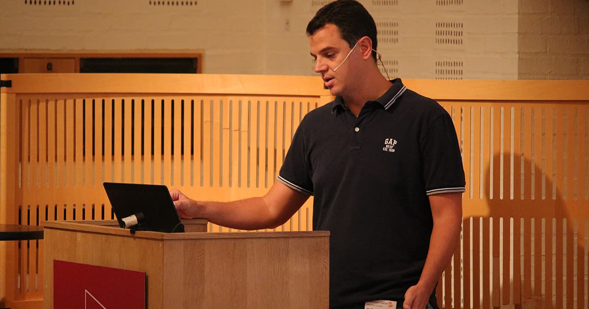 Francesco Garue of SimCorp Italiana stands at a podium delivering a talk at Dyalog ’19. He is wearing a dark GAP polo shirt and speaking into a headset microphone, while interacting with a laptop. The setting includes a wooden podium and slatted wood backdrop.