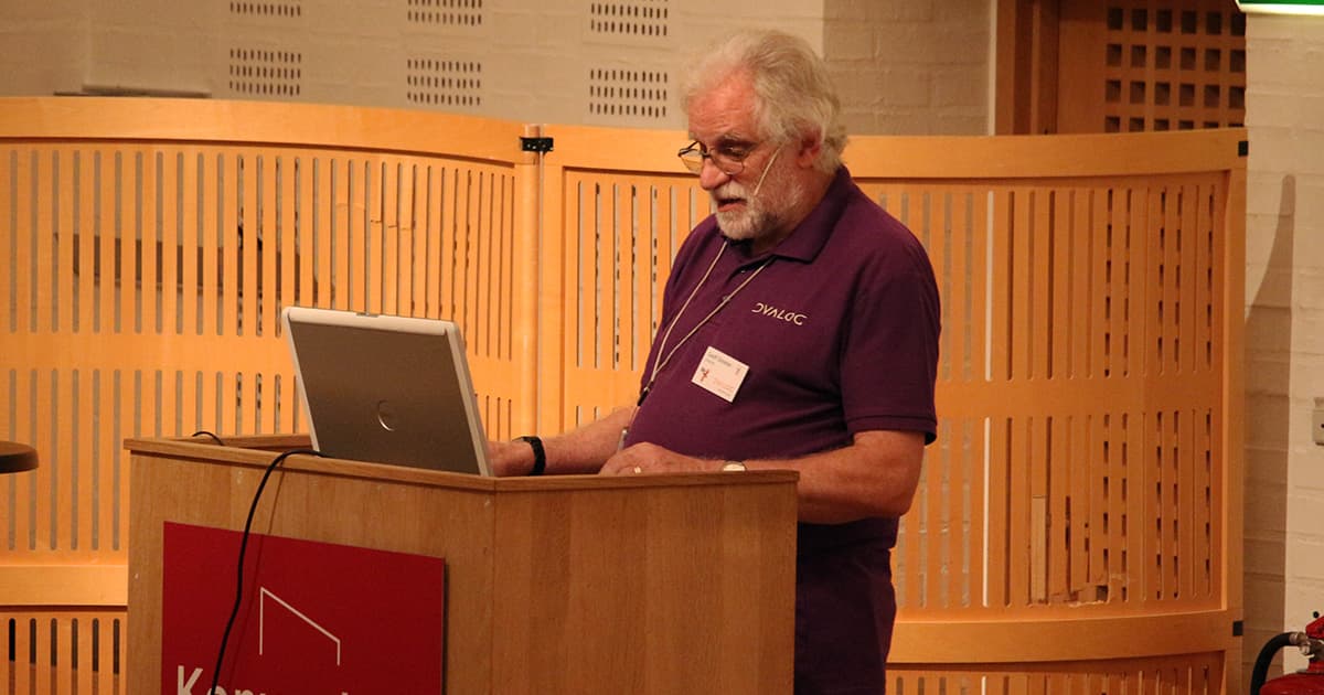 Geoff Streeter of Dyalog Ltd speaks at a podium during Dyalog ’19. He is wearing a purple Dyalog polo shirt and headset microphone, looking at a laptop while discussing the history of shared code files. The background features a light wood panel design.