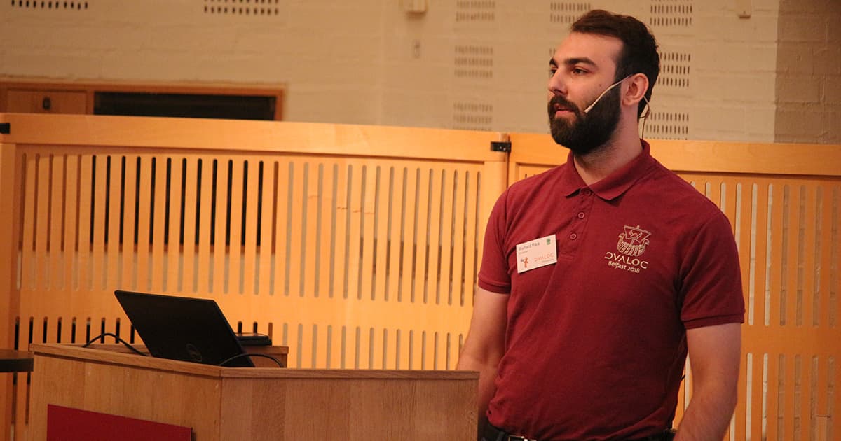 Richard Park presents TamStat at Dyalog ’19. He stands beside a podium, wearing a headset microphone and a maroon Dyalog Belfast 2018 polo shirt, in a wood-panelled lecture room at Konventum.