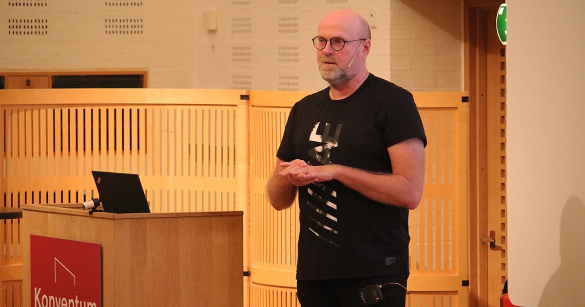 Claus Madsen of FinE Analytics speaks at Dyalog ’19. He stands beside a podium, wearing a black t-shirt and headset mic, with hands clasped as he addresses the audience in a wood-panelled lecture hall at Konventum.