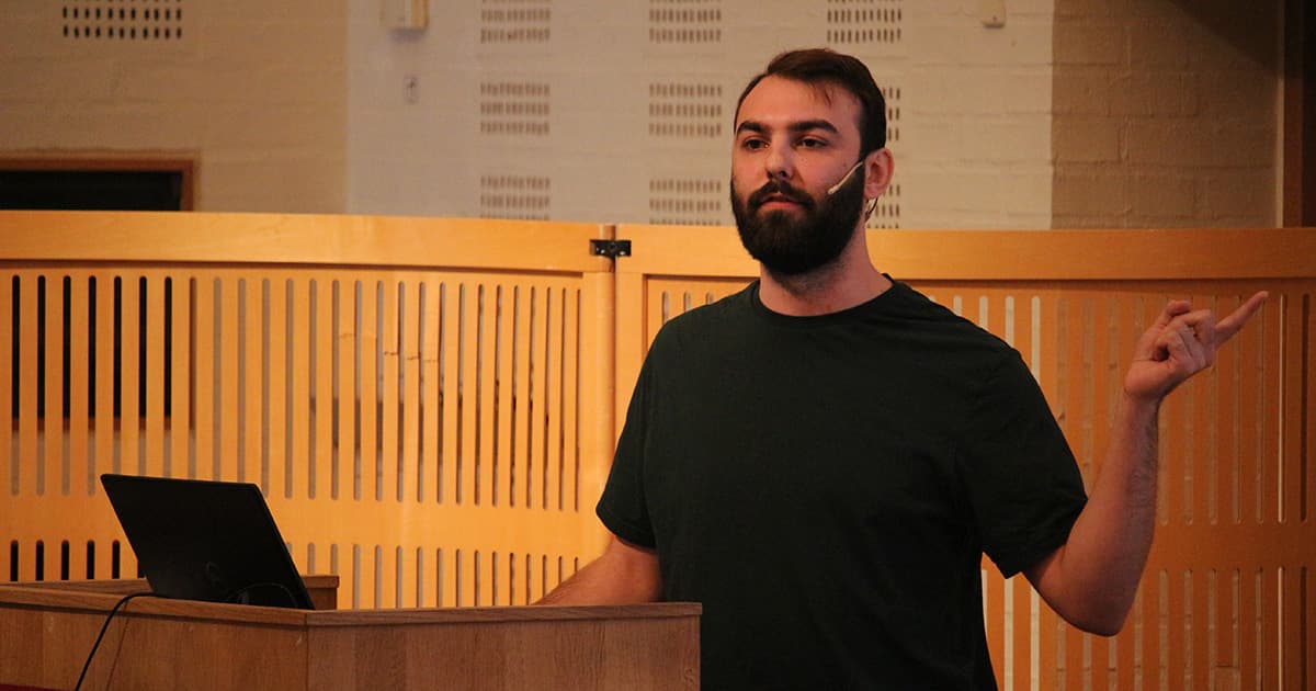 Rich Park presenting at Dyalog ’19. He stands beside a wooden podium, gesturing with one hand while wearing a headset microphone and a dark green T-shirt, in a wood-panelled lecture hall at Konventum.