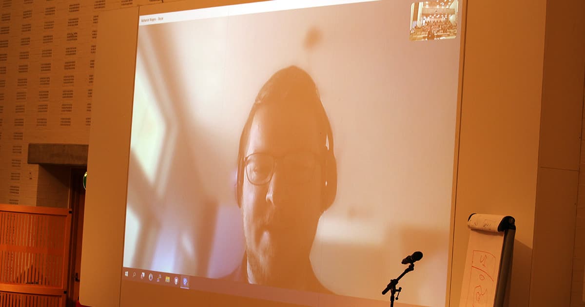 A live video call projected onto a large screen in a conference hall shows Nathan Rogers, wearing glasses and a headset, presenting remotely. The screen includes a small thumbnail of the in-person audience in the corner. A microphone and flip chart are visible in front of the projection.