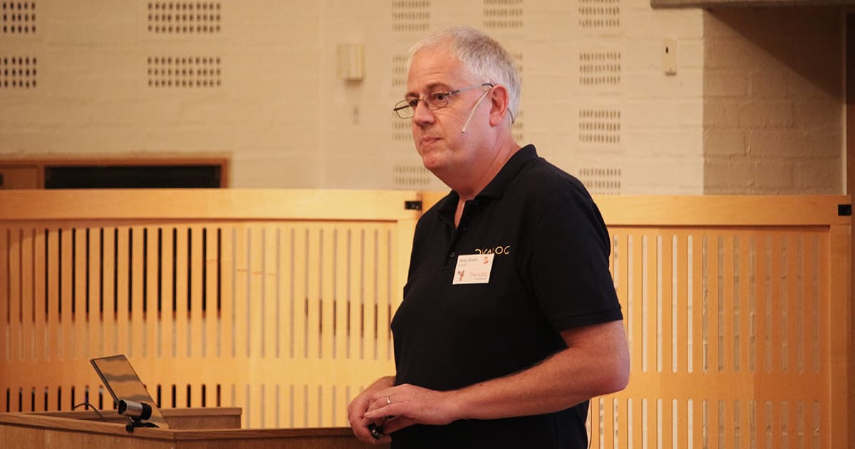Andy Shiers, wearing a Dyalog-branded polo shirt and a headset microphone, stands mid-speech during his informal “fireside chat” session at Dyalog ’19. He has grey hair and is holding a small object in his hands, with the wooden panel backdrop of the Konventum venue behind him.