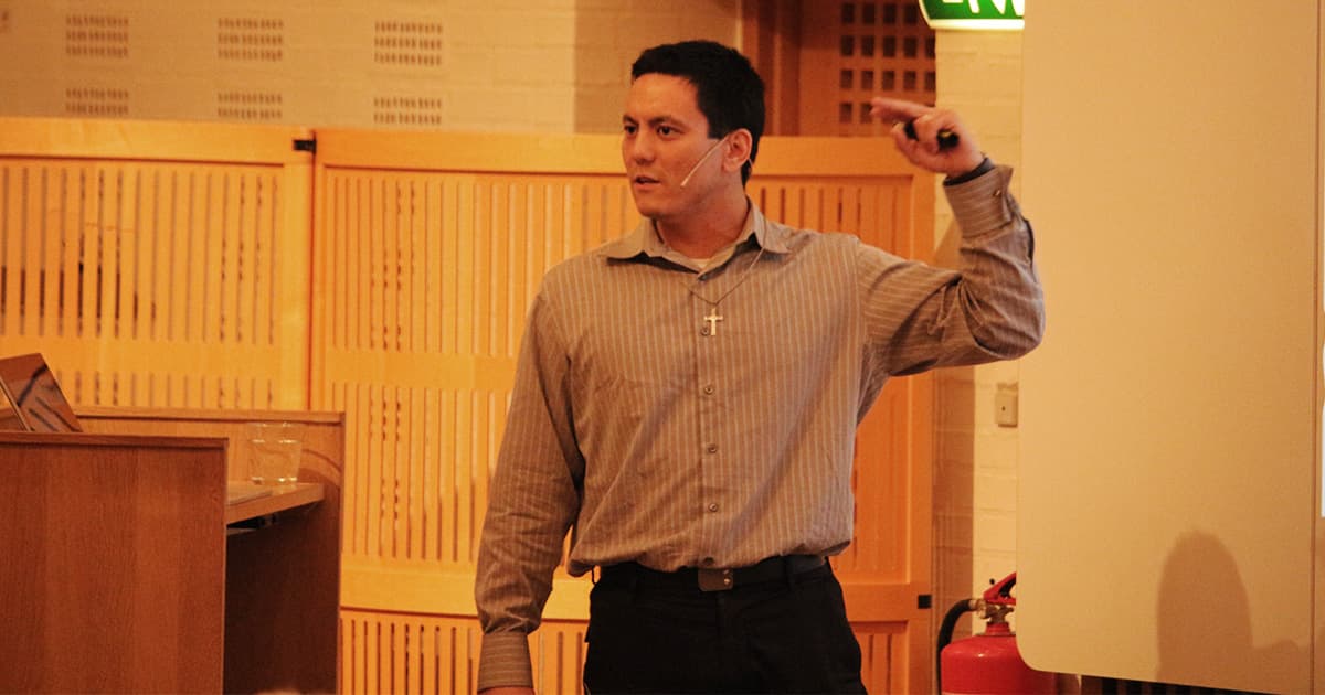Aaron Hsu stands on stage mid-presentation, wearing a headset and striped shirt with a cross necklace. He gestures to a screen out of view while speaking about his work on the Co-dfns APL compiler project. A laptop, glasses of water, and fire extinguisher are visible nearby in the conference setting.