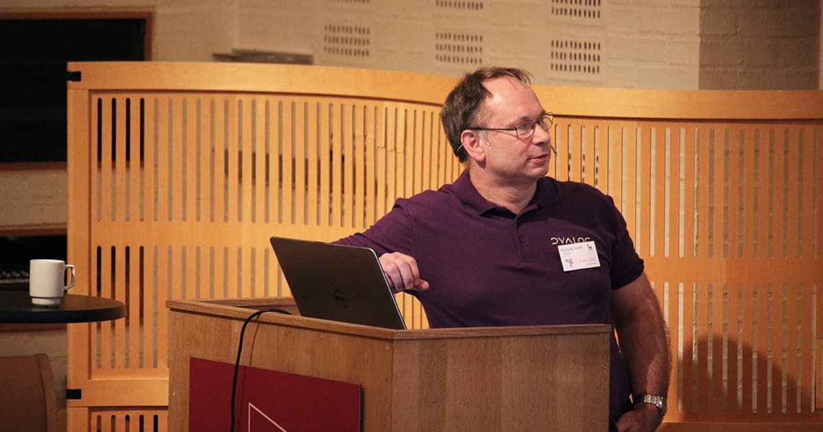 Richard Smith stands behind a lectern during Dyalog ’19, wearing a Dyalog-branded polo shirt and headset microphone. He gestures slightly while looking to his left, with a laptop open in front of him and a coffee mug visible on a small round table. The wood-panelled backdrop of the Konventum venue frames the scene.