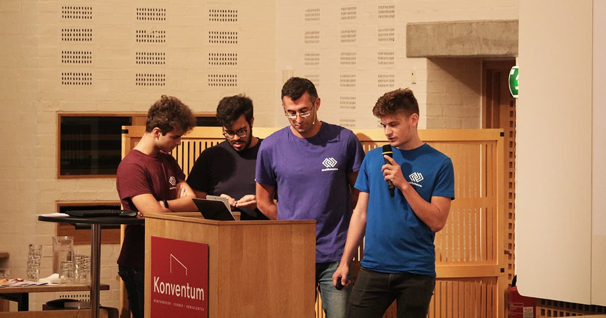 Roberto and three students from Liceo Scientifico GB Grassi Saronno present at Dyalog ’19. One student speaks into a microphone while using a tablet, with the others standing attentively nearby. All are wearing MathsBeyond shirts.
