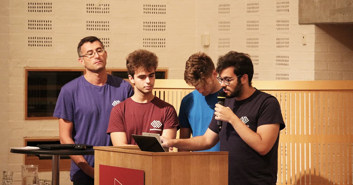 Roberto and three students from Liceo Scientifico GB Grassi Saronno present at Dyalog ’19. One student speaks into a microphone while using a tablet, with the others standing attentively nearby. All are wearing MathsBeyond shirts.