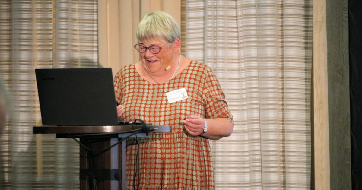 Gitte Christensen, Managing Director of Dyalog Ltd, smiling as she welcomes attendees to the Dyalog '22 user meeting in Olhão, Portugal, standing at a podium with a laptop.