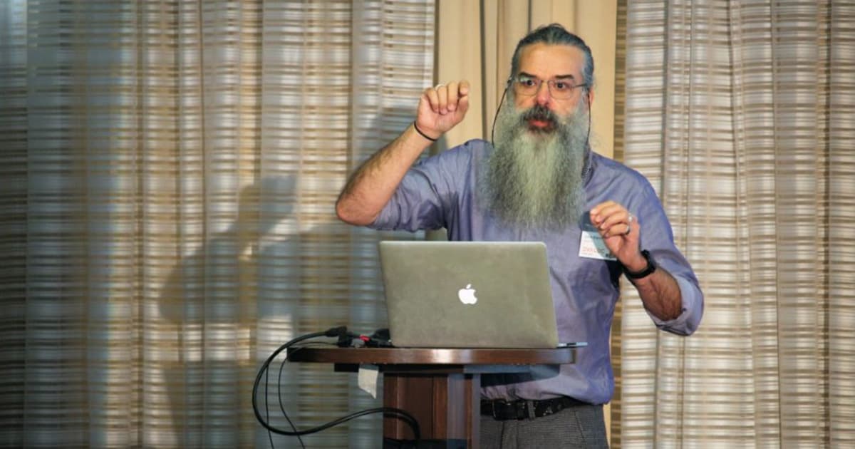 John Daintree presenting at Dyalog '22 in Olhão, Portugal, sharing ideas about asynchronous programming while gesturing expressively behind a podium with a MacBook.
