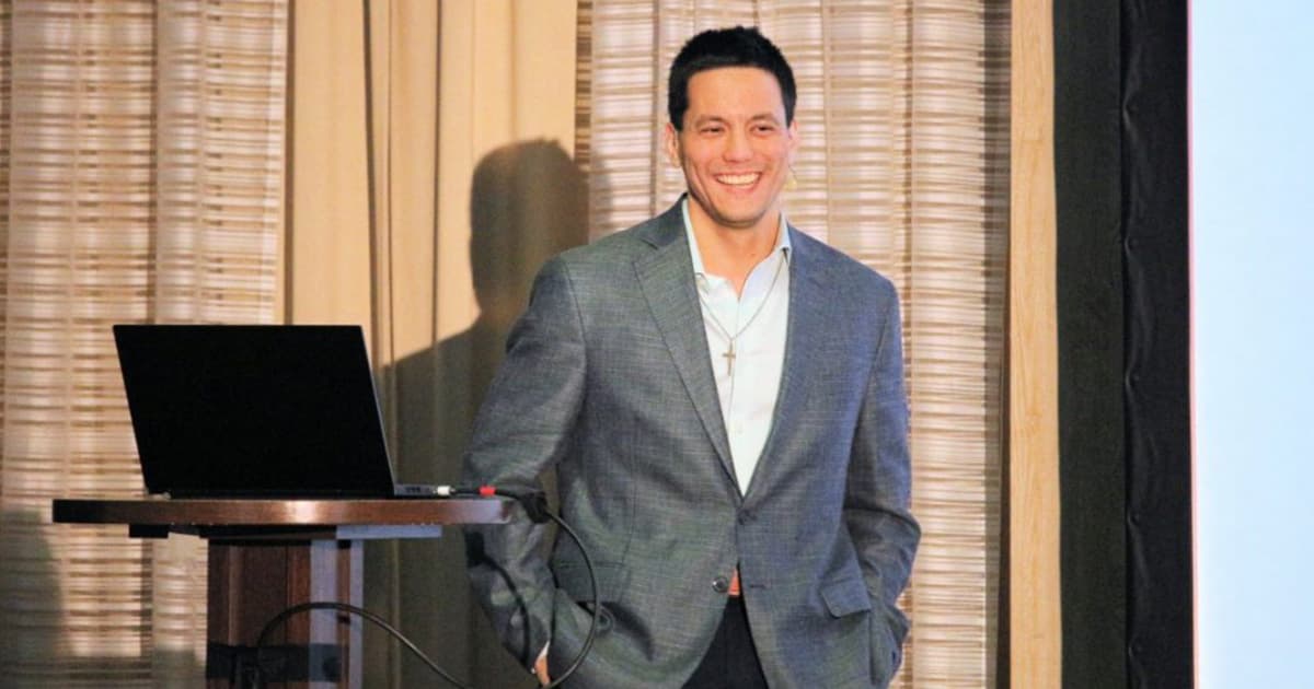 Aaron Hsu presenting at Dyalog '22 in Olhão, Portugal, smiling as he delivers an update on the Co-dfns compiler, standing beside a podium with a laptop.