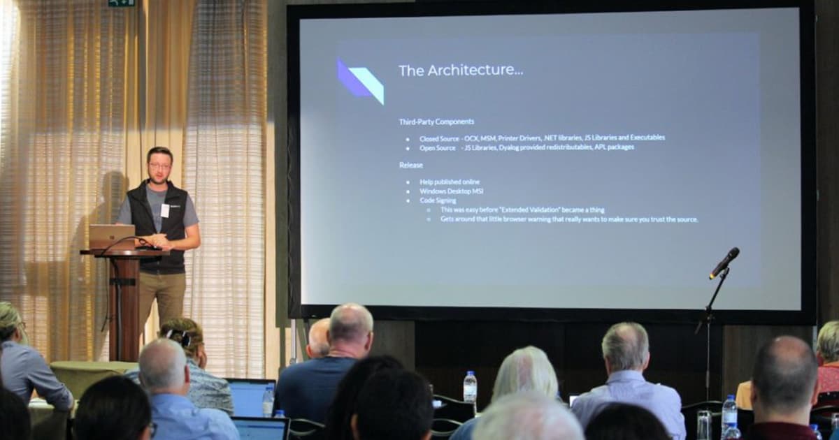 Norbert Jurkiewicz presenting at Dyalog '22 in Olhão, Portugal, discussing automation architecture while standing beside a large screen displaying a slide on third-party components and deployment strategy.