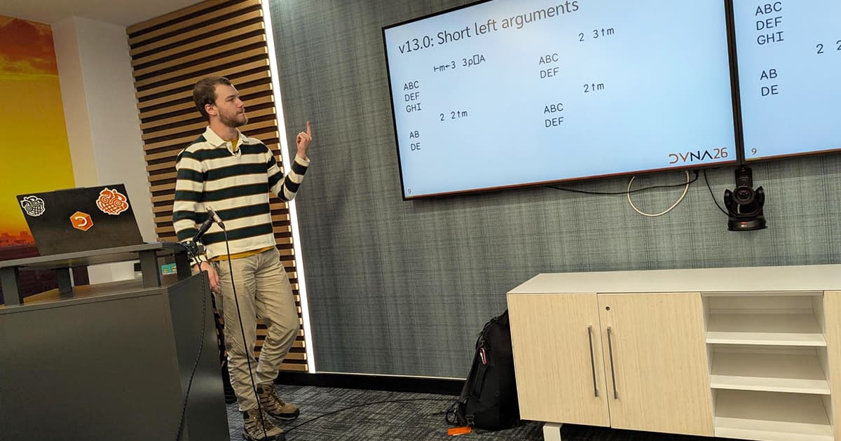 Asher Harvey-Smith presenting at DYNA26, standing beside screens showing APL examples and a slide titled “v13.0: Short left arguments.”