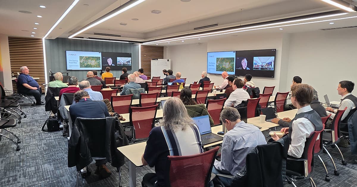 Attendees at DYNA26 seated in a conference room watching a presentation, with slides and a remote speaker shown on screens at the front.