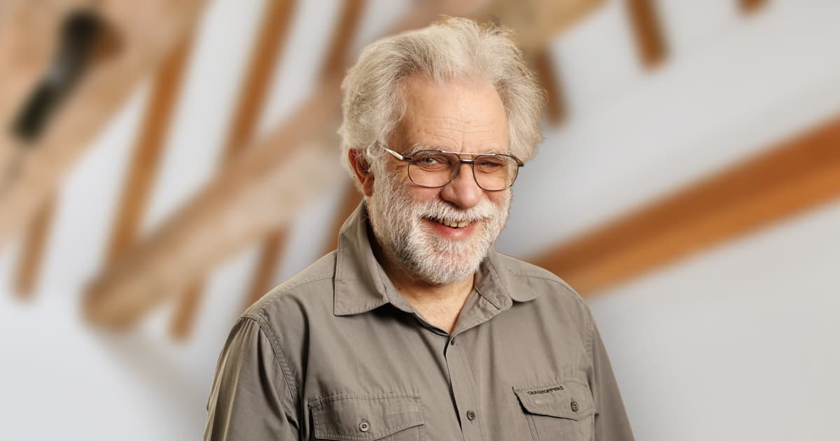 Geoff Streeter smiles warmly in front of a softly blurred architectural wooden beam background, wearing a grey shirt and glasses.