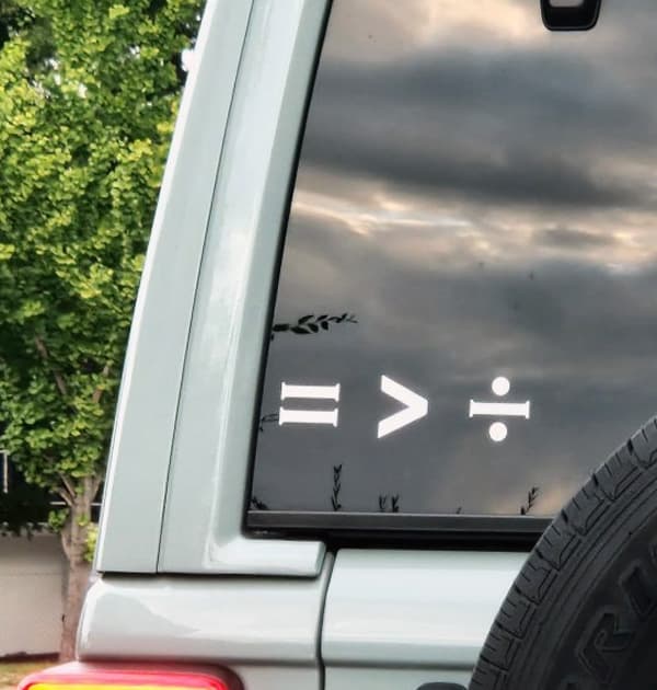 White APL symbols displayed as stickers on the rear window of a vehicle: equal sign, greater-than sign, and division sign.