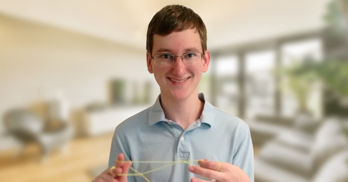 Marshall Lochbaum smiles while holding a cat’s cradle string figure, standing in front of a softly blurred modern living room.