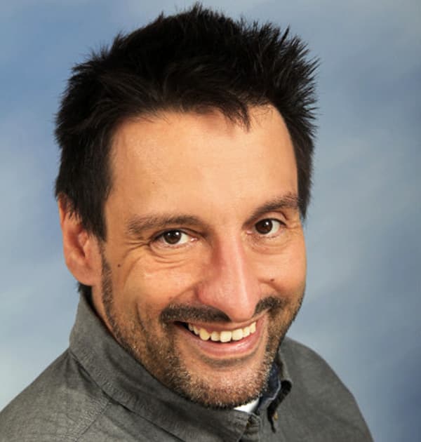 Michael Baas smiles at the camera in front of a soft blue studio background, wearing a grey button-up shirt.