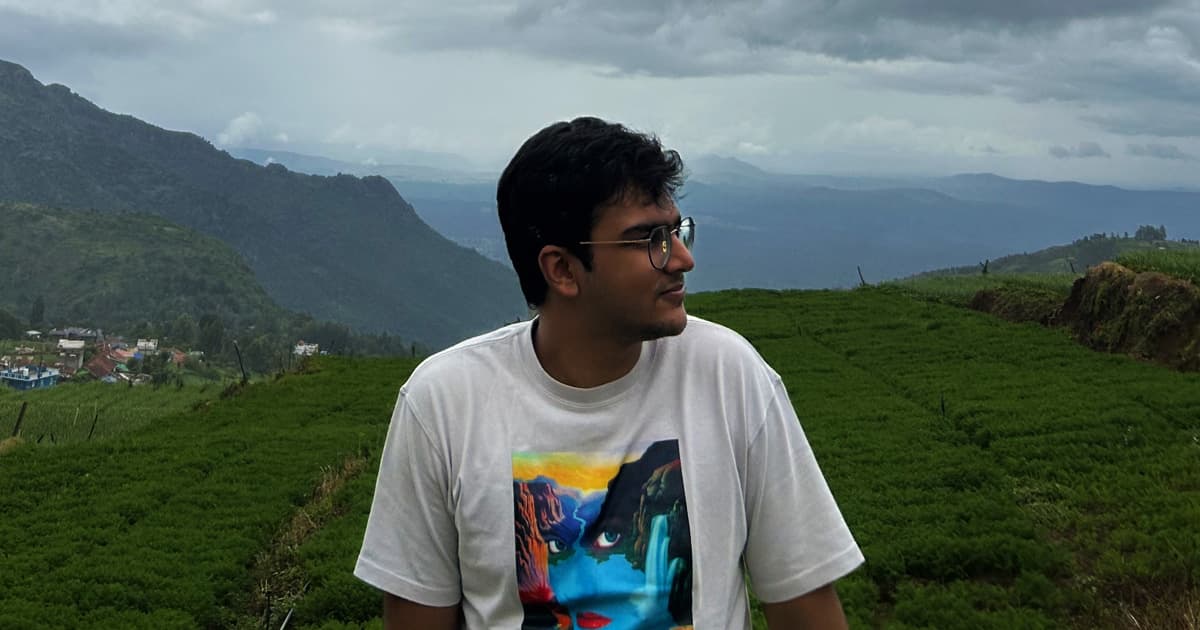 Aarush Bhat sitting in a lush green tea plantation with rolling hills and a misty, overcast sky in the background. He is wearing glasses and a white T-shirt featuring a colourful abstract face design, looking off to the side.