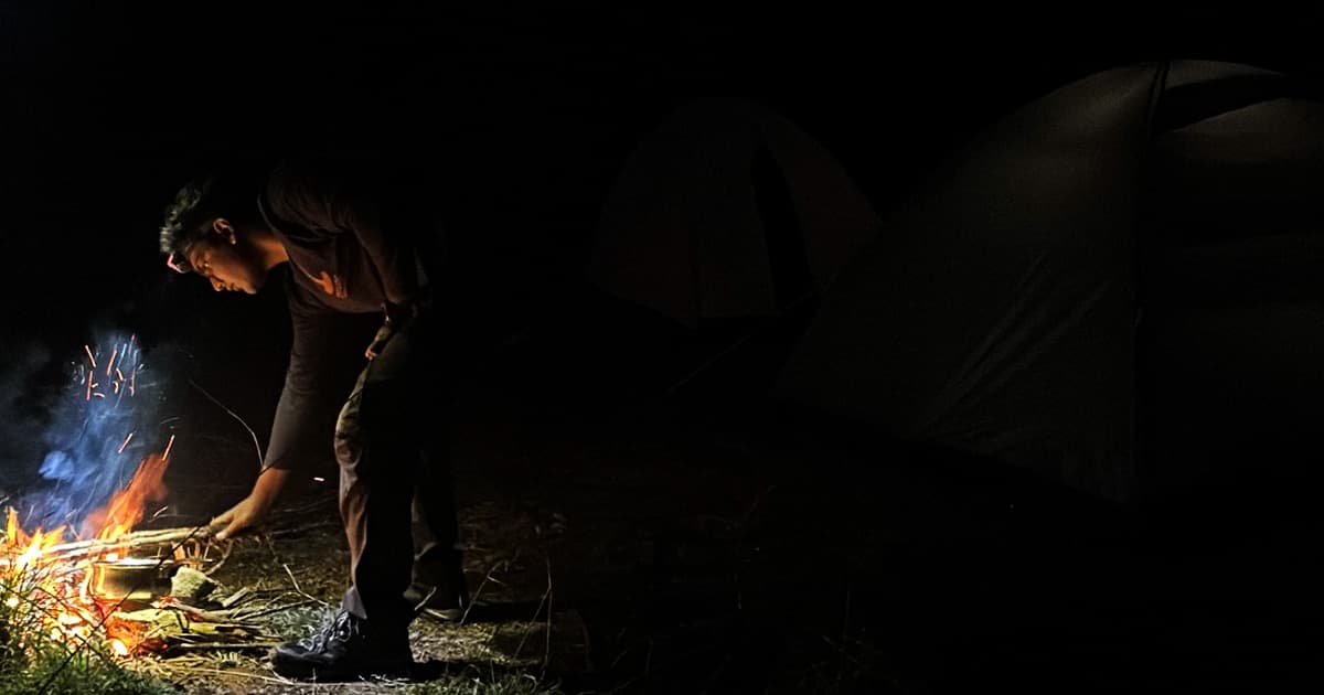 Aarush Bhat tending to a small campfire at night, wearing a headlamp and dark clothing. Sparks and smoke rise from the fire as he adjusts a pot over the flames. Two tents are visible in the background, dimly lit by the fire's glow.