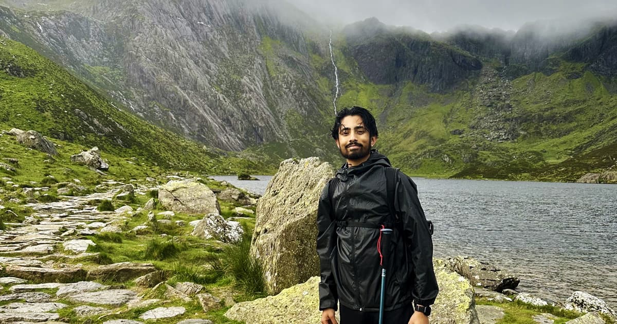 Abs Suri standing on a rocky trail beside a mountain lake, wearing a black waterproof jacket and carrying a hiking pole. Behind him, a waterfall cascades down a mist-covered mountain, surrounded by lush green slopes. The cloudy sky and damp terrain suggest recent rainfall.