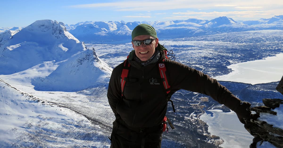 Stefan Kruger smiling at the summit of a snowy mountain with panoramic views of Norway’s rugged winter landscape.