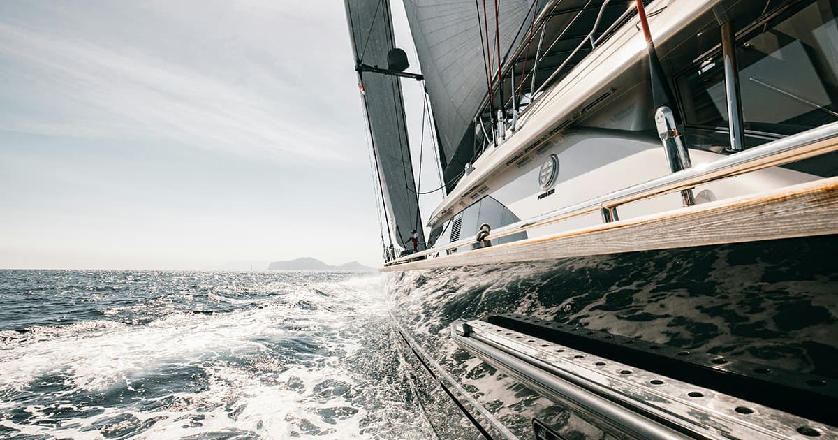 A close-up side view of a sleek sailing yacht cutting through the water at speed, with sunlight reflecting off the waves and distant islands on the horizon.