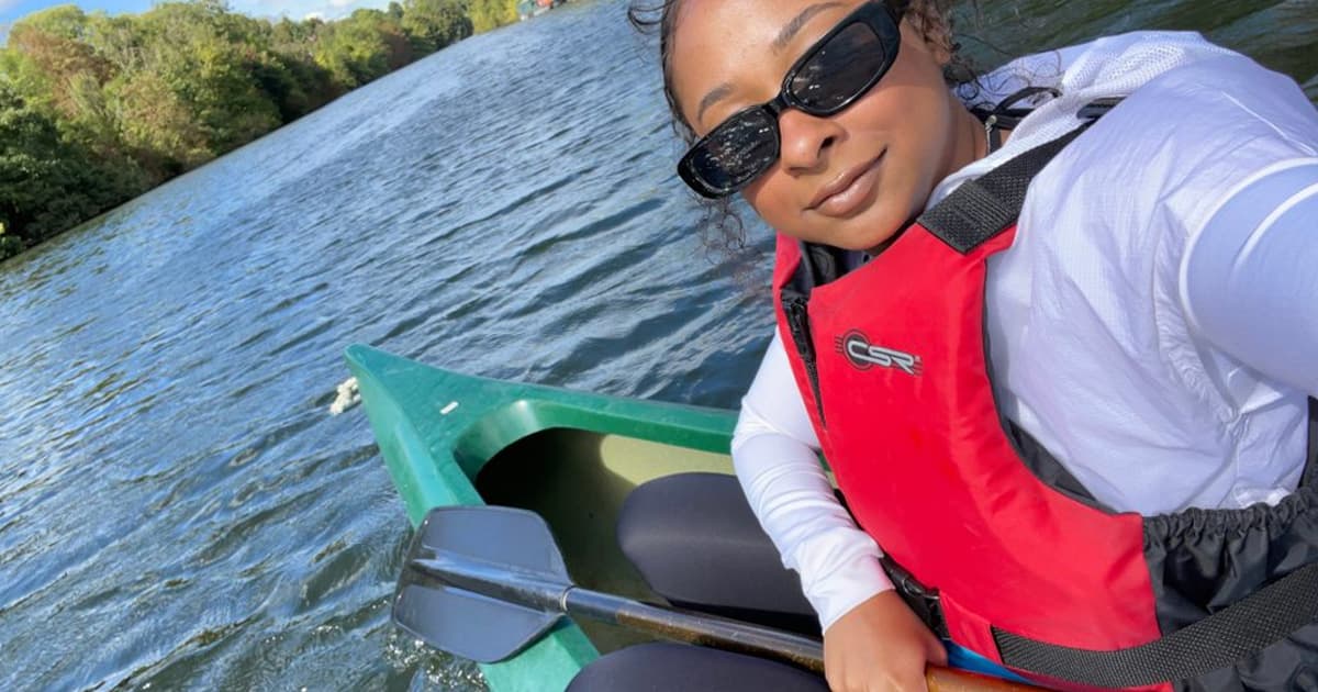 Jada Andrade smiles while sitting in a green canoe on a river, wearing a red life jacket, sunglasses, and holding a paddle, surrounded by calm water and trees.