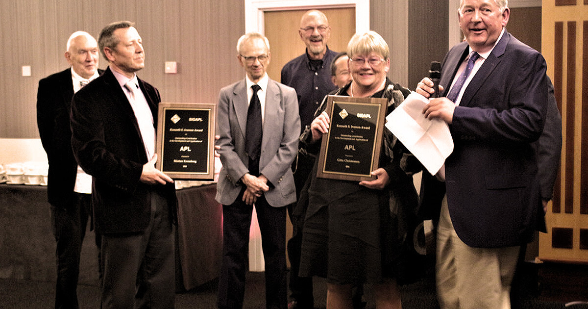 Morten Kromberg and Gitte Christensen receive the 2016 Iverson Award at a formal event. Gitte holds a plaque while Paul Grosvenor of Optima Systems Ltd speaks into a microphone.