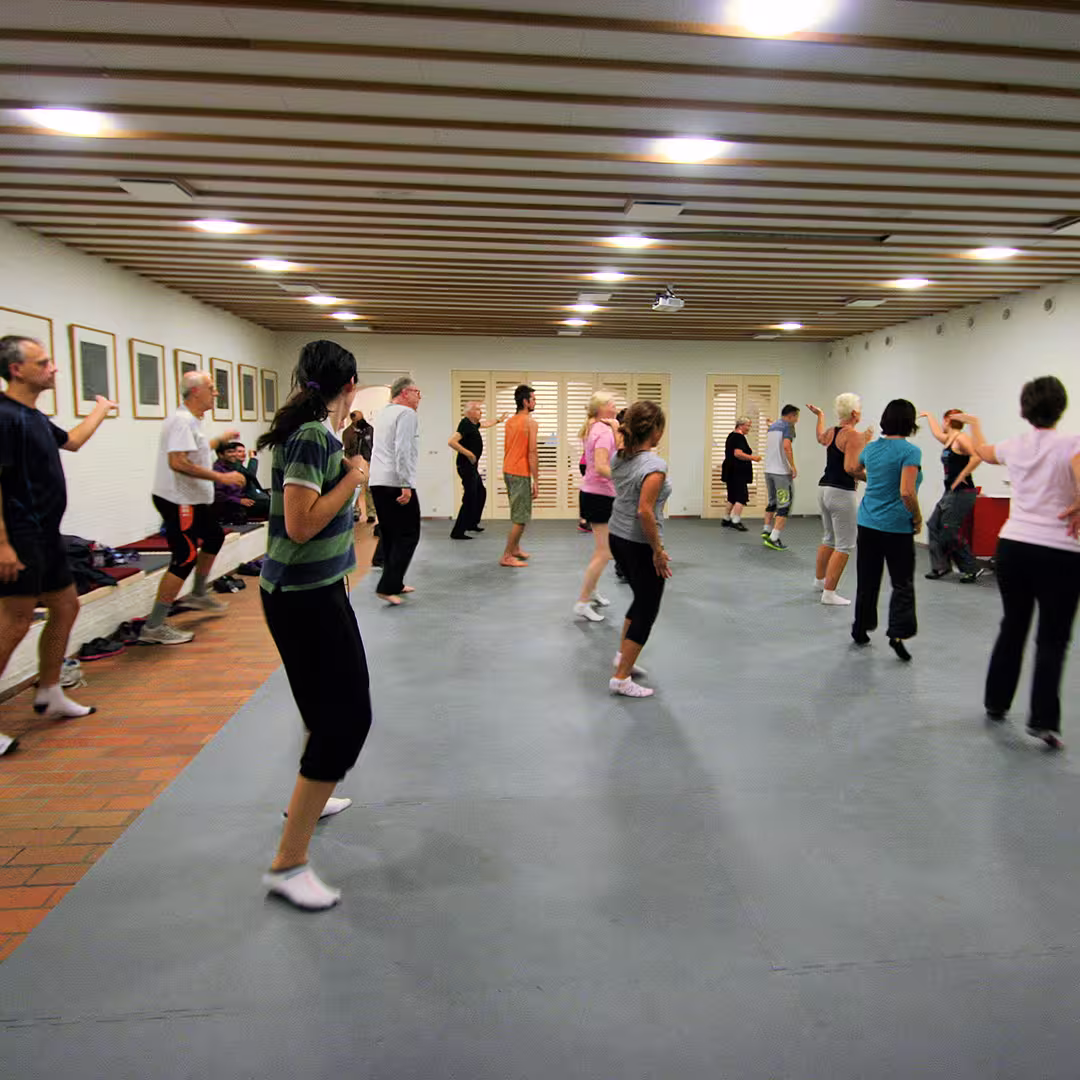 A group of Dyalog '12 delegates participating in a group exercise session at the Konventum Conference Centre. They are dressed in casual athletic wear and following an instructor's lead in a spacious room with wooden ceiling beams and framed artwork on the walls.