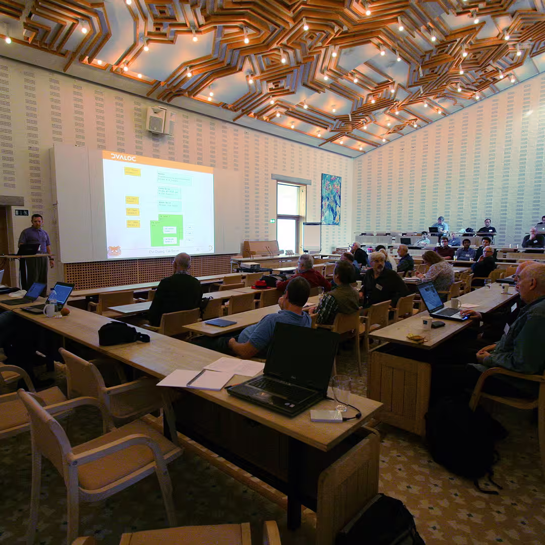 Morten Kromberg presenting at the Dyalog '12 event in a conference hall at the Konventum Conference Centre. He is standing at the front, next to a large screen displaying a flowchart. Delegates are seated at wooden desks, some using laptops. The room features a striking geometric ceiling design with embedded lights and a colorful abstract painting on the wall.