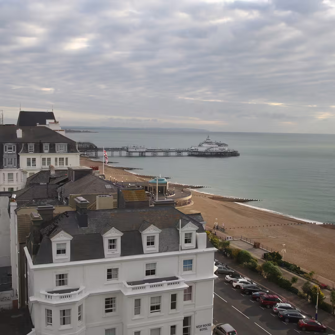 View of Eastbourne seafront, UK, showing the West Rocks Hotel in the foreground, the sandy beach, and the Eastbourne Pier extending into the calm sea under a cloudy sky during the Dyalog '14 event.