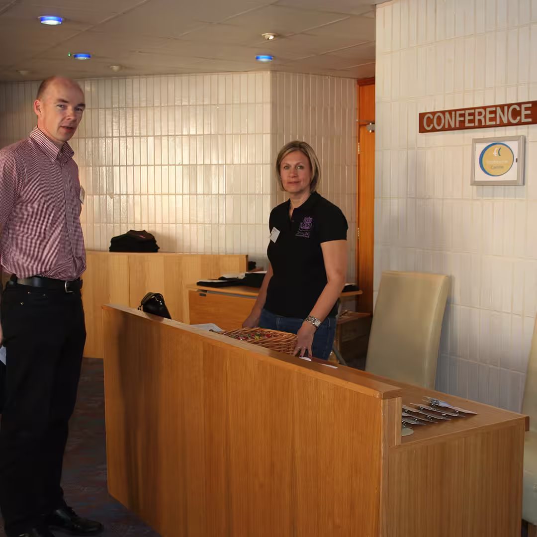 Karen Shaw of Dyalog Ltd standing at the registration desk during the Dyalog '14 event in Eastbourne, UK. She is speaking with a delegate near the conference entrance, with event materials and a basket of sweets on the desk.