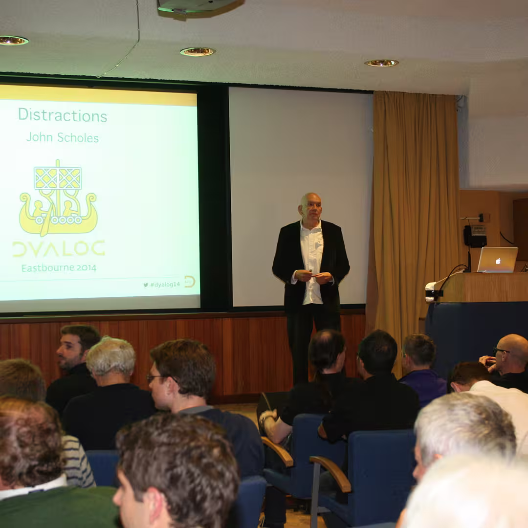 John Scholes presenting a talk titled "Distractions" at the Dyalog '14 event in Eastbourne, UK. He stands in front of a screen displaying the Dyalog logo and event details, with an audience seated in blue chairs.