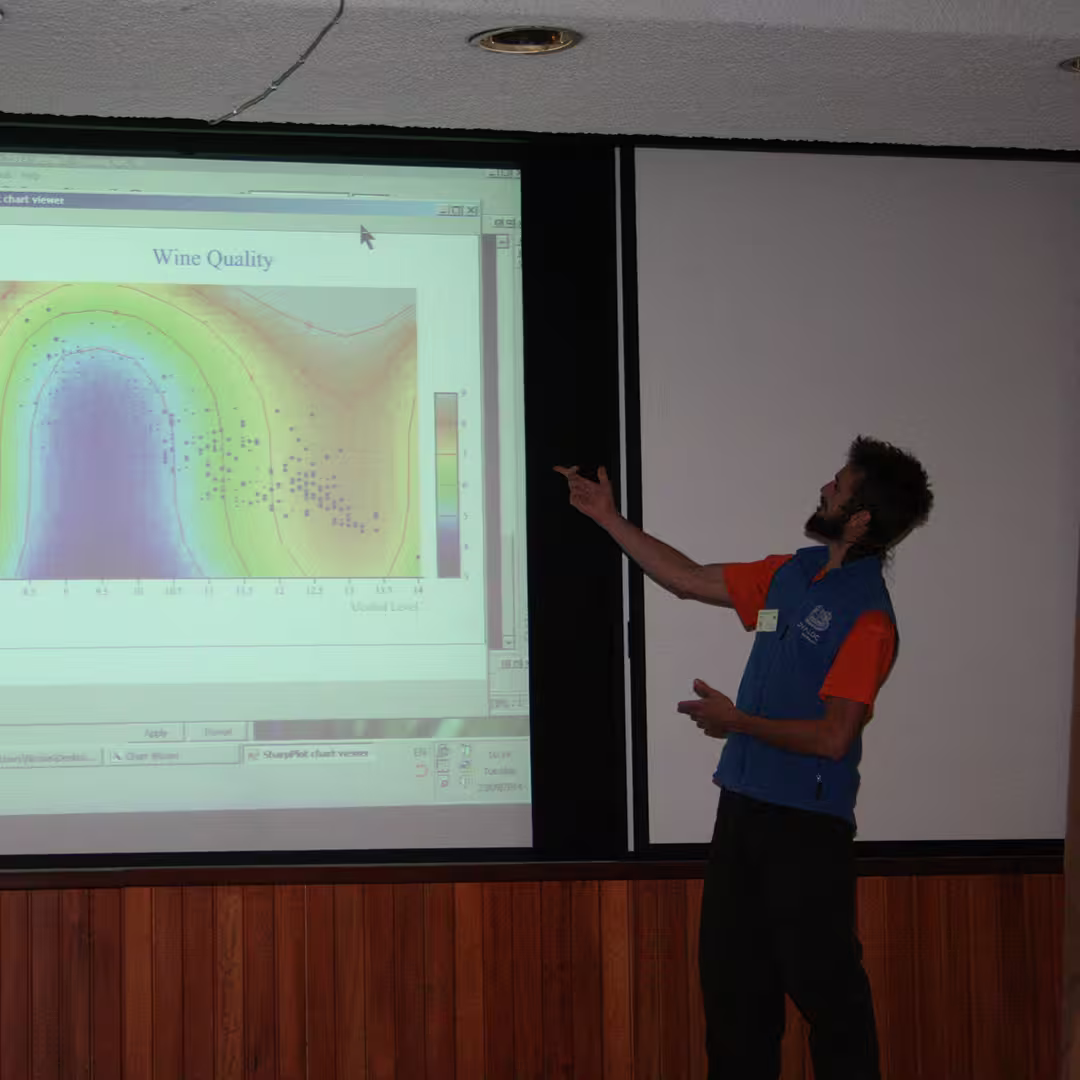 A Dyalog '14 delegate presenting a slide titled "Wine Quality" in Eastbourne, UK. He is pointing to a colorful contour plot on the screen, showing the relationship between alcohol level and wine quality.