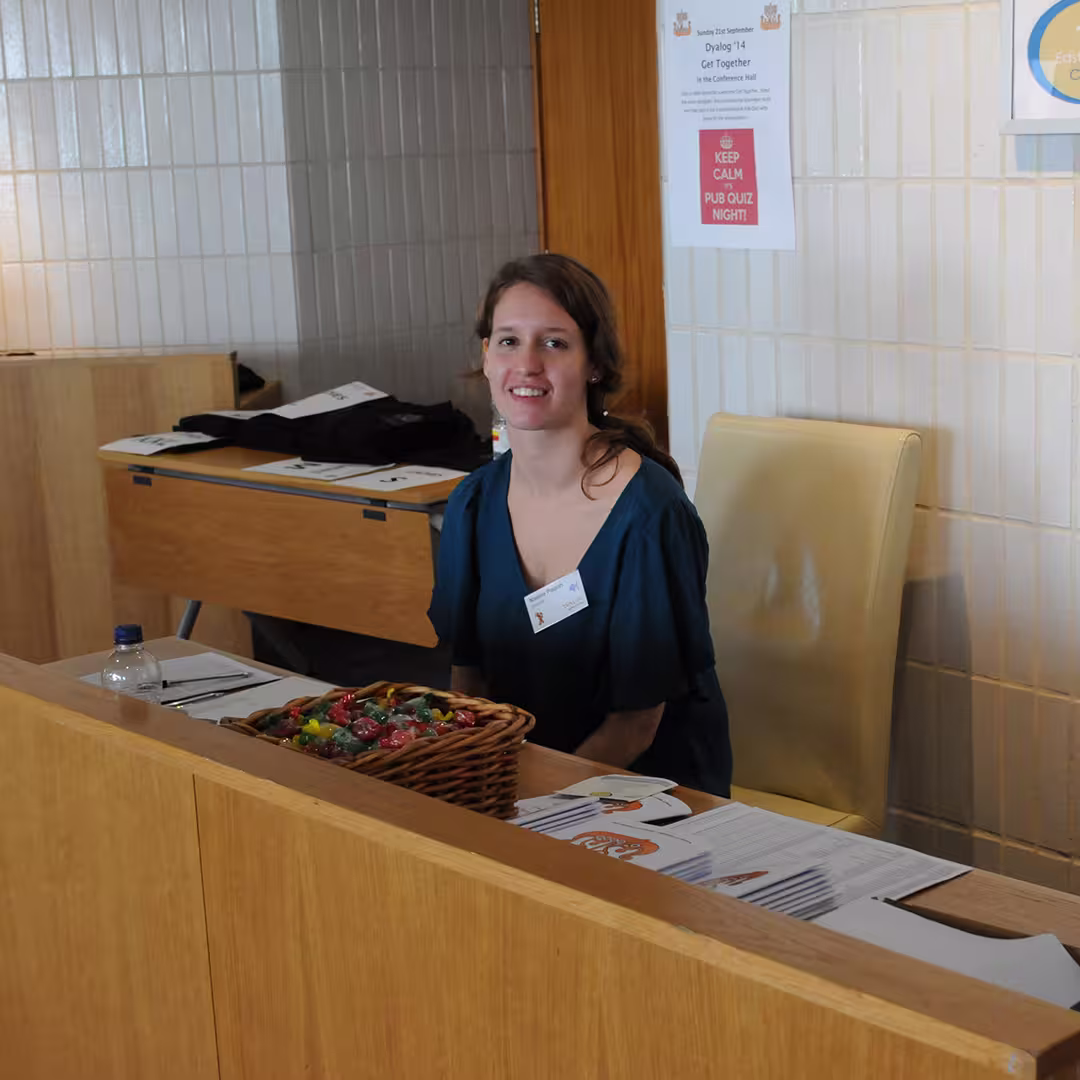 A Dyalog Ltd staff memnber seated at the registration desk in Eastbourne, UK. The desk has event materials, a basket of sweets, and a water bottle. Behind her, a sign reads "Keep Calm Pub Quiz Night!"
