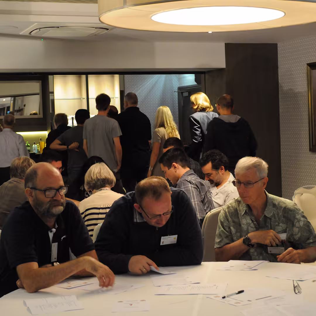 Dyalog '14 delegates seated at a table, focusing on documents during an evening event in Eastbourne, UK. Other attendees are seen standing near the bar in the background, engaged in conversation.