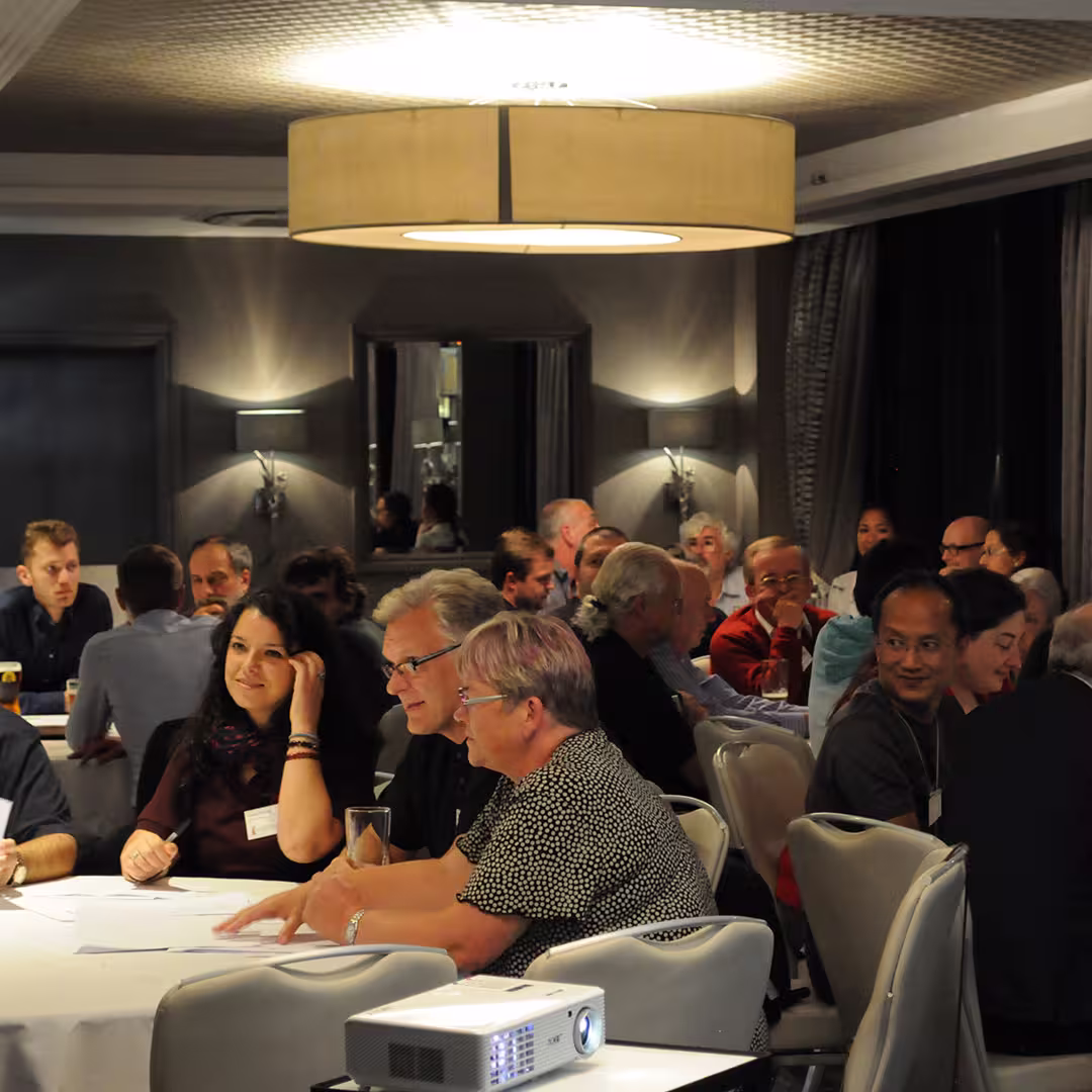 Dyalog '14 delegates seated around tables during an evening event in Eastbourne, UK. Attendees are engaged in conversation, with papers, drinks, and a projector visible on the table. The room is softly lit with modern décor.